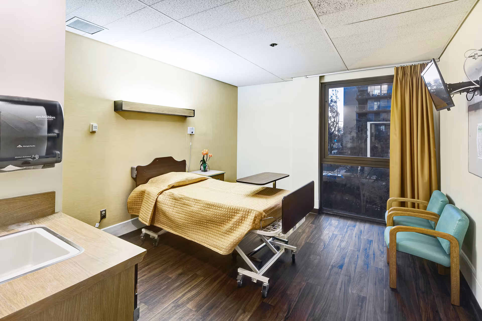Private resident room with a single adjustable bed, bedside table, sink area, two chairs and a wall-mounted TV by a large window.