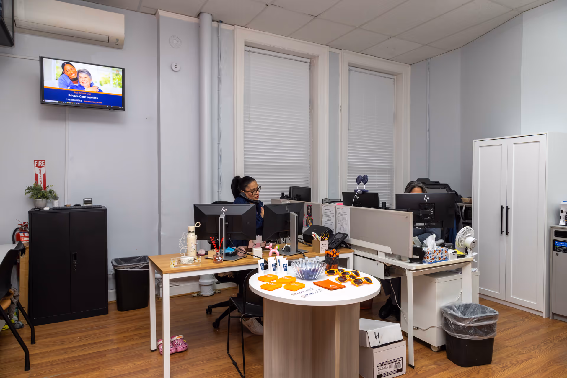 An office workspace with two staff at computers, desks, and office equipment in a light-colored room.