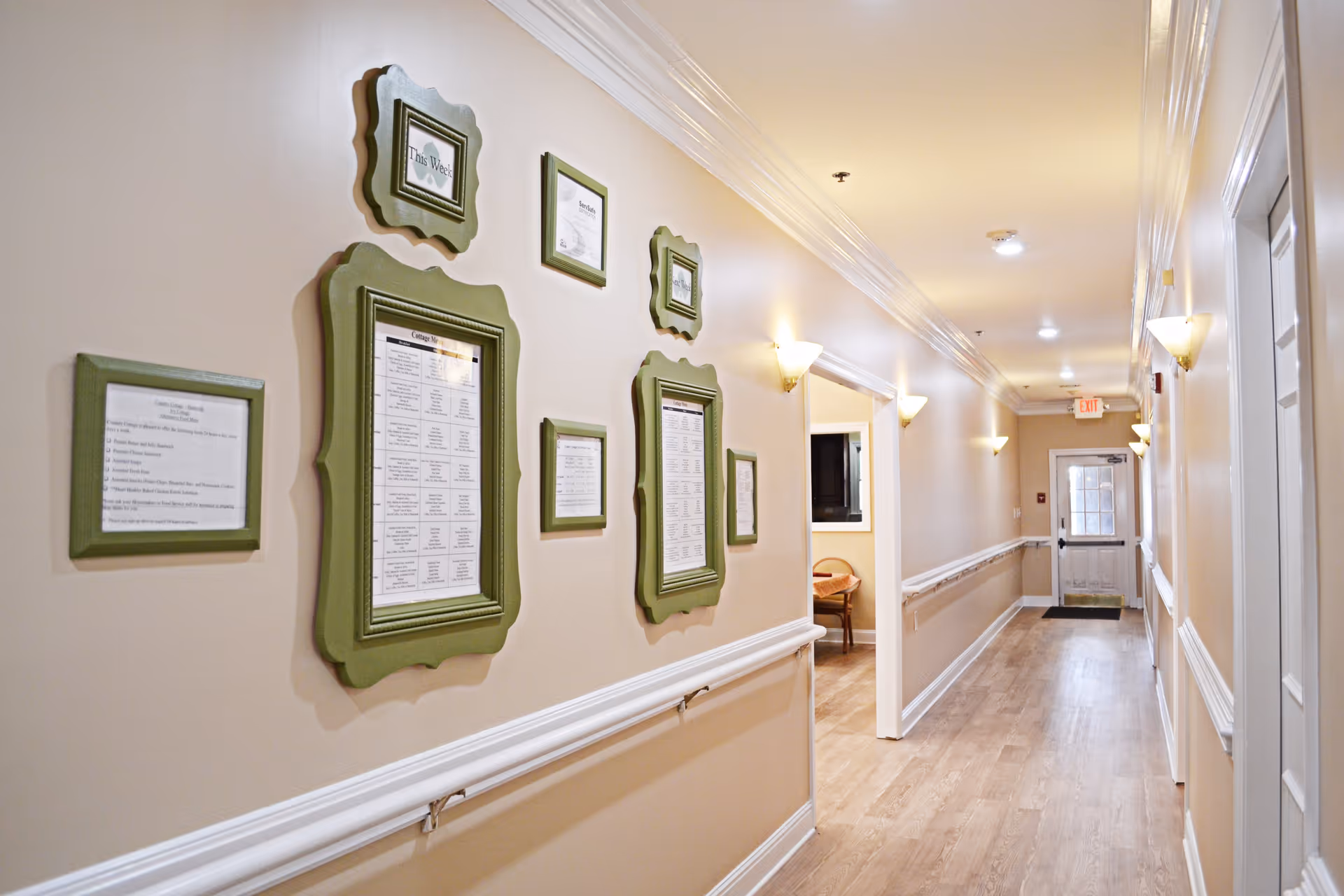 A well-lit hallway in a senior living facility with light-colored walls and wood flooring. The left wall features several green-framed notices and schedules. Handrails run along both sides of the hallway. There are wall sconces providing warm lighting and a door with a window at the far end.