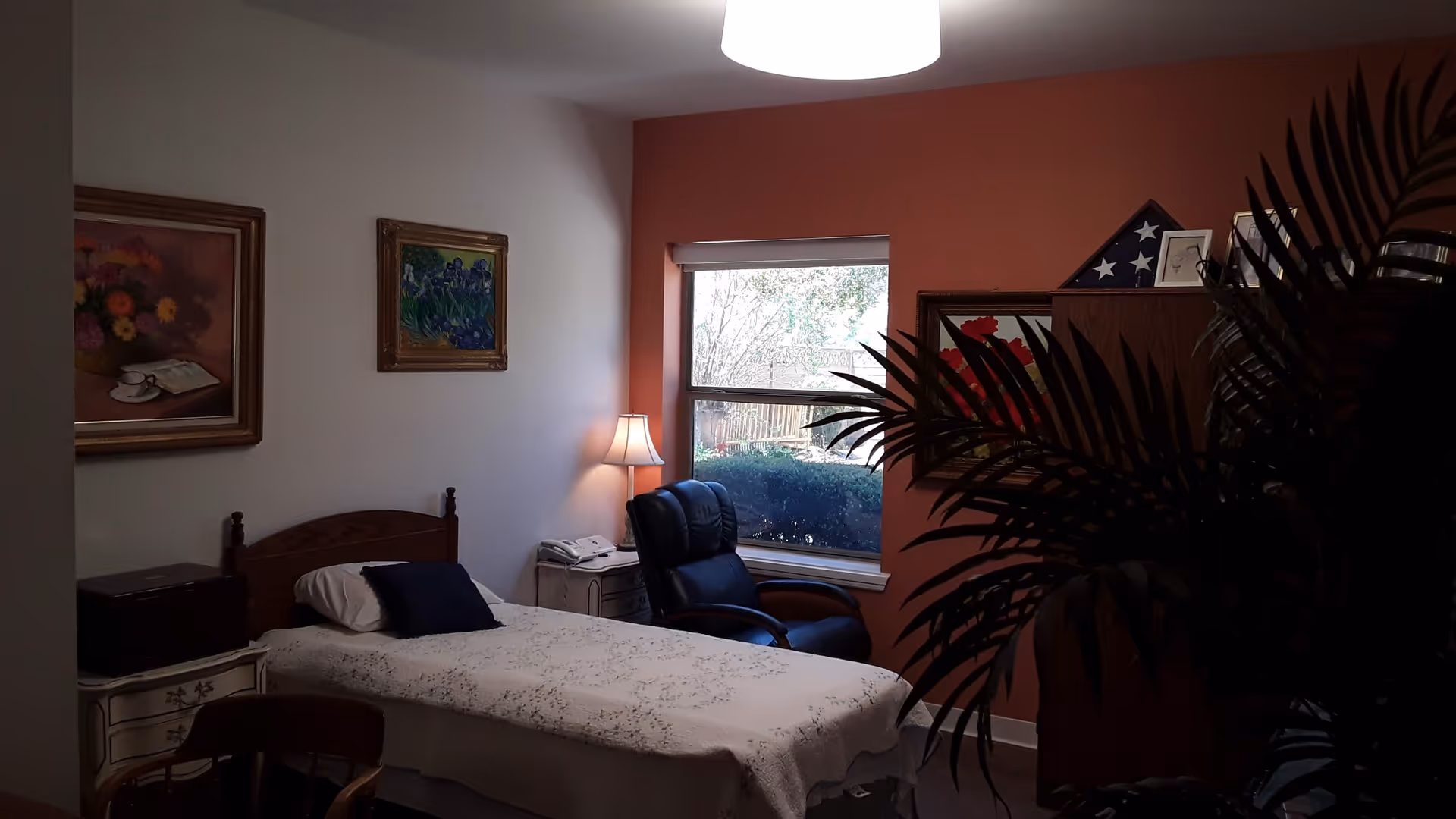 A cozy bedroom with bed, nightstand, lamp, recliner near window, framed paintings, plant.