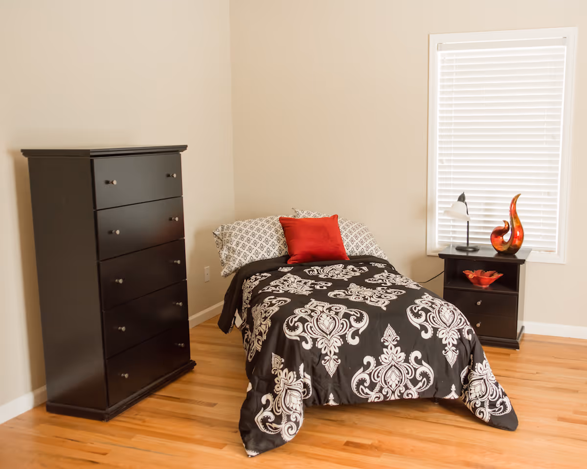 A bedroom with a single bed covered in a black and white patterned comforter and a red pillow. Next to the bed is a black nightstand with a lamp and decorative items. There is a tall black dresser against the wall and a window with white blinds. The floor is wooden and the walls are light-colored.