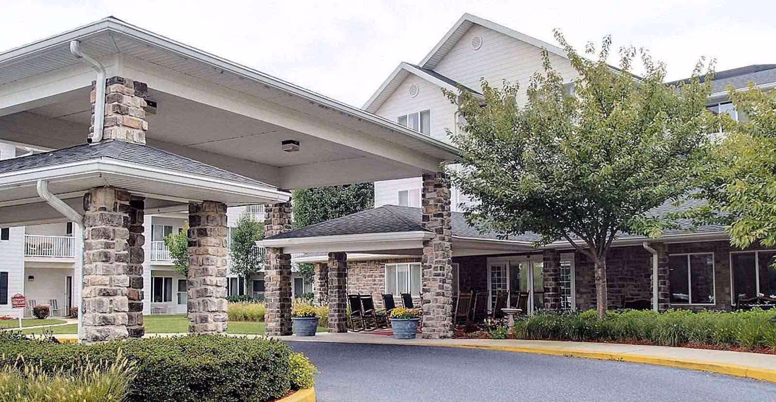 Exterior view of a senior living facility entrance with stone pillars supporting a covered driveway, surrounded by greenery and trees.