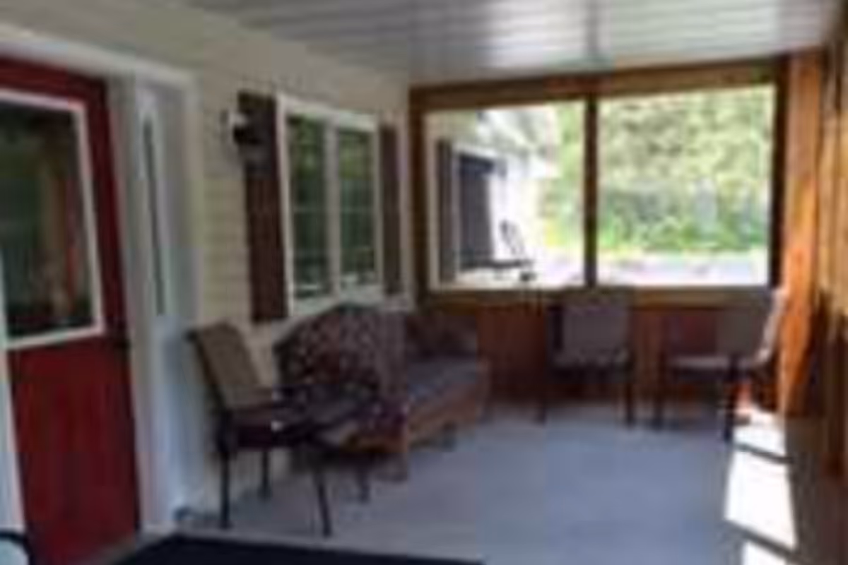 Sunlit enclosed porch with chairs, a sofa, and large windows overlooking greenery outside.
