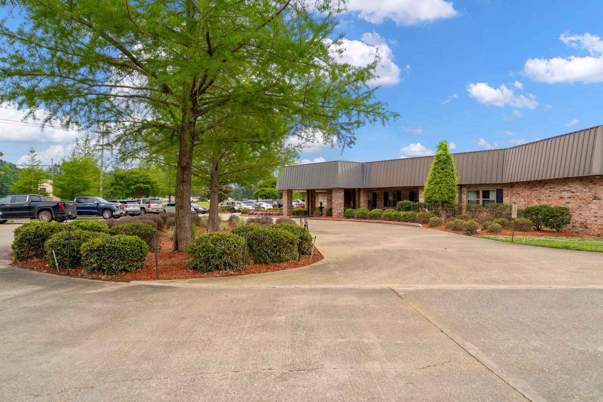 Exterior view of a single-story brick building with a metal roof, surrounded by landscaped bushes and trees. A curved driveway leads to the entrance, and several parked cars are visible in the background under a partly cloudy sky.