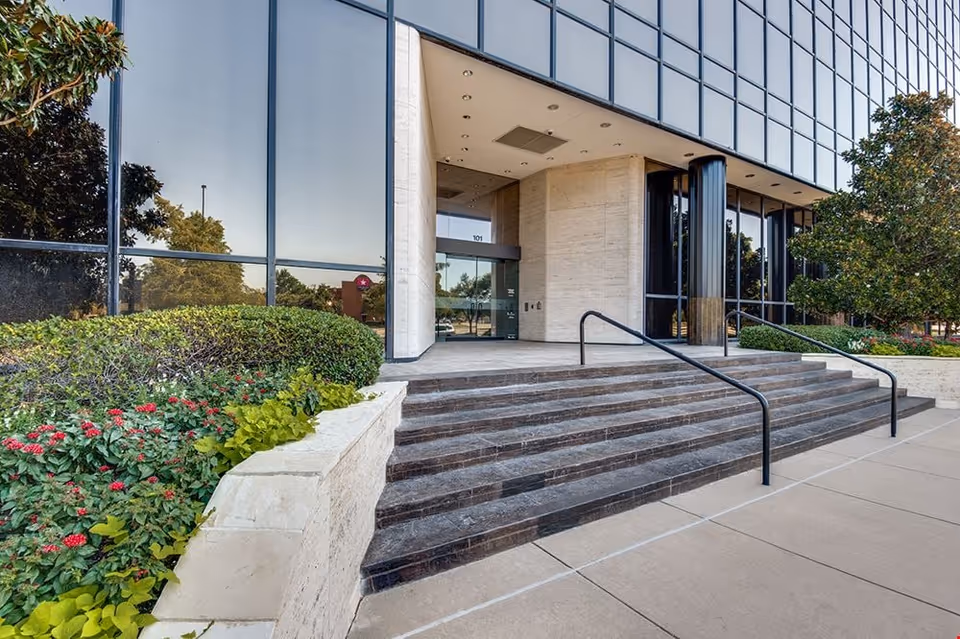 Front entrance of a modern glass-building with steps and landscaped shrubs.