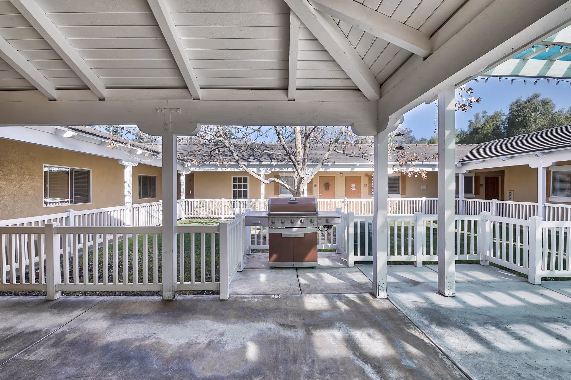 Covered outdoor patio area with a barbecue grill in the center, surrounded by white fencing. The patio overlooks a grassy courtyard with a tree in the middle, and single-story building units with windows and doors encircle the courtyard.