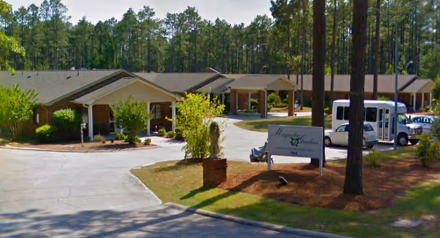 Single story Magnolia Gardens senior living facility with driveway entrance and surrounding pine trees.