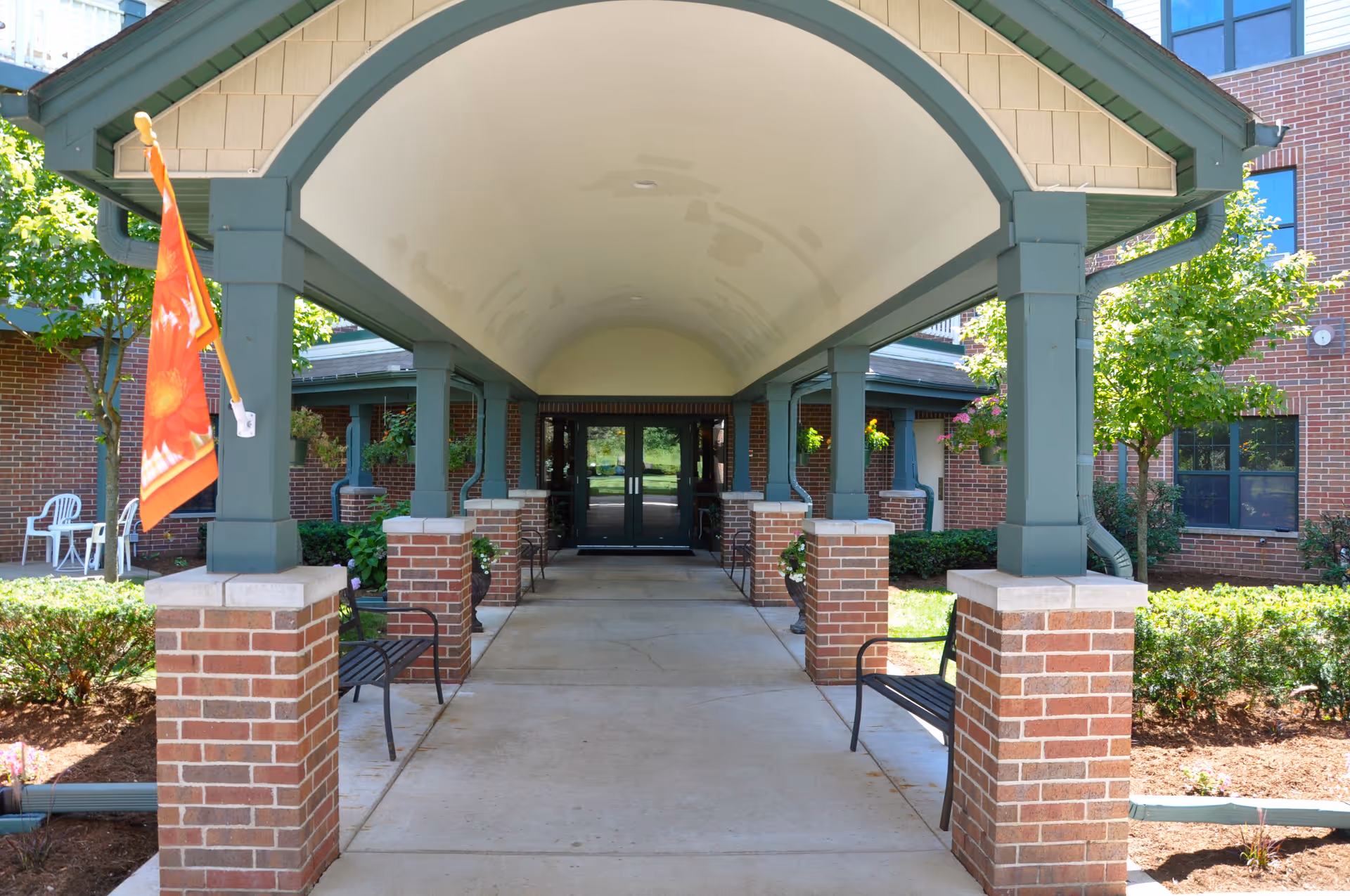 Covered walkway entrance to a brick building with green pillars and benches on either side, surrounded by landscaped bushes and trees, with a bright orange flag on the left pillar.