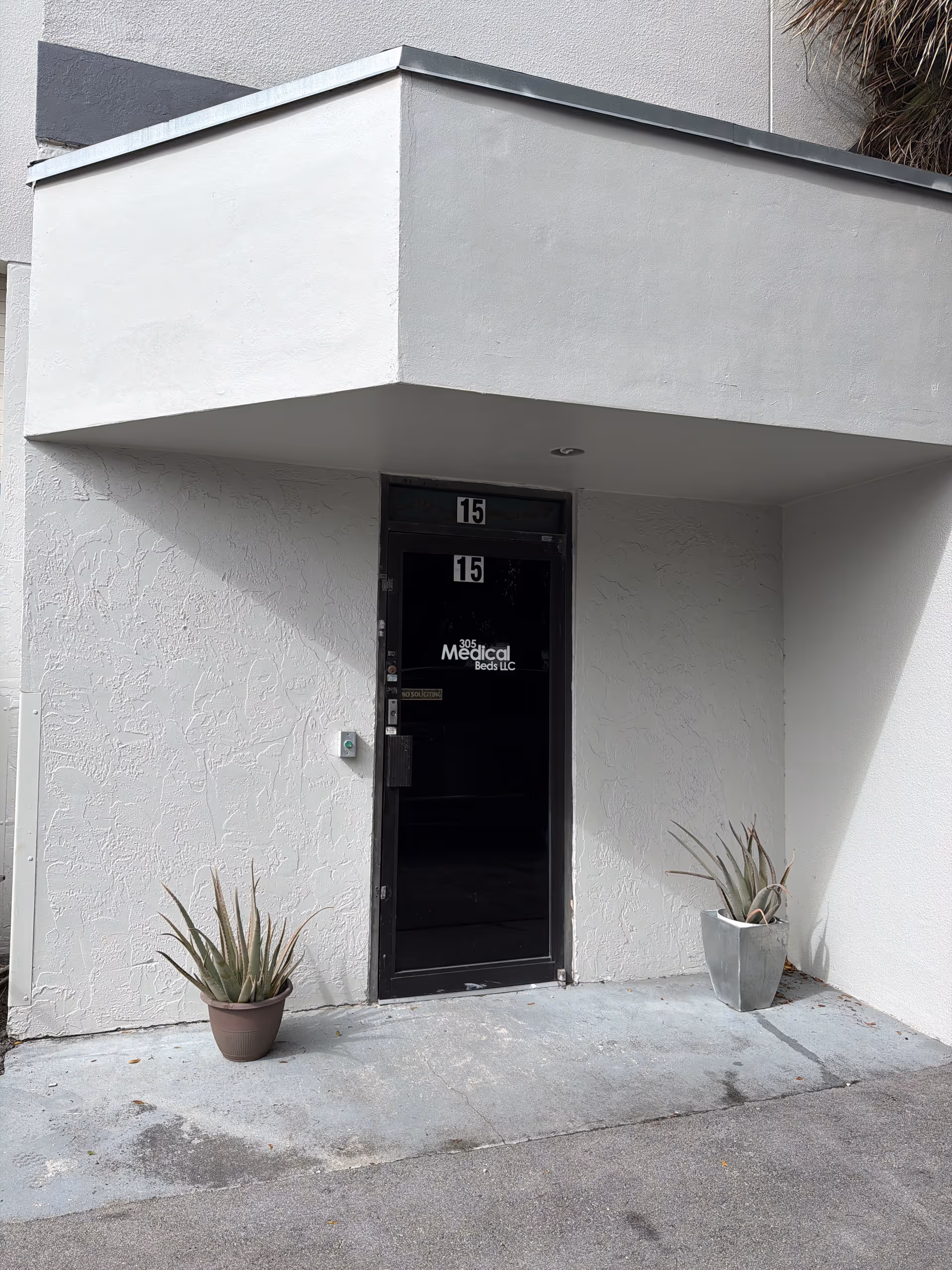 Exterior building entrance with a black door labeled 305 Medical Beds LLC and two potted plants.