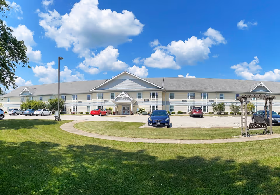 Front exterior view of a two-story senior living facility building with a gray roof and light-colored siding under a bright blue sky with scattered clouds. There is a parking lot with several cars in front of the building and a curved sidewalk leading to the entrance. A wooden bench with a pergola is visible on the right side of the image, and green grass and trees surround the area.