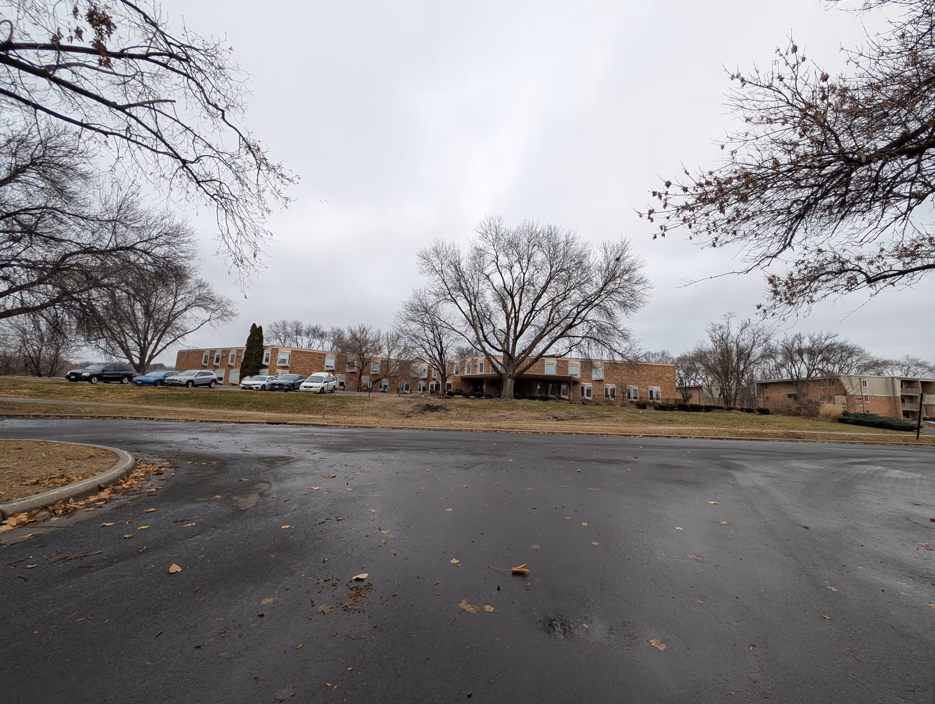 Low-angle view of a brick senior living building with parked cars, leafless trees, and wet pavement under an overcast sky.