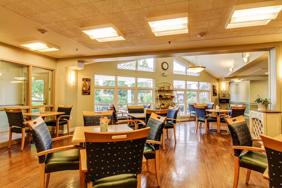 A bright dining room with multiple wooden tables and chairs arranged neatly. The room has large windows allowing natural light to fill the space, with a view of greenery outside. The ceiling has recessed lighting, and there are decorative elements such as a clock, wall art, and a small shelving unit. The floor is wooden, and the overall atmosphere is warm and inviting.