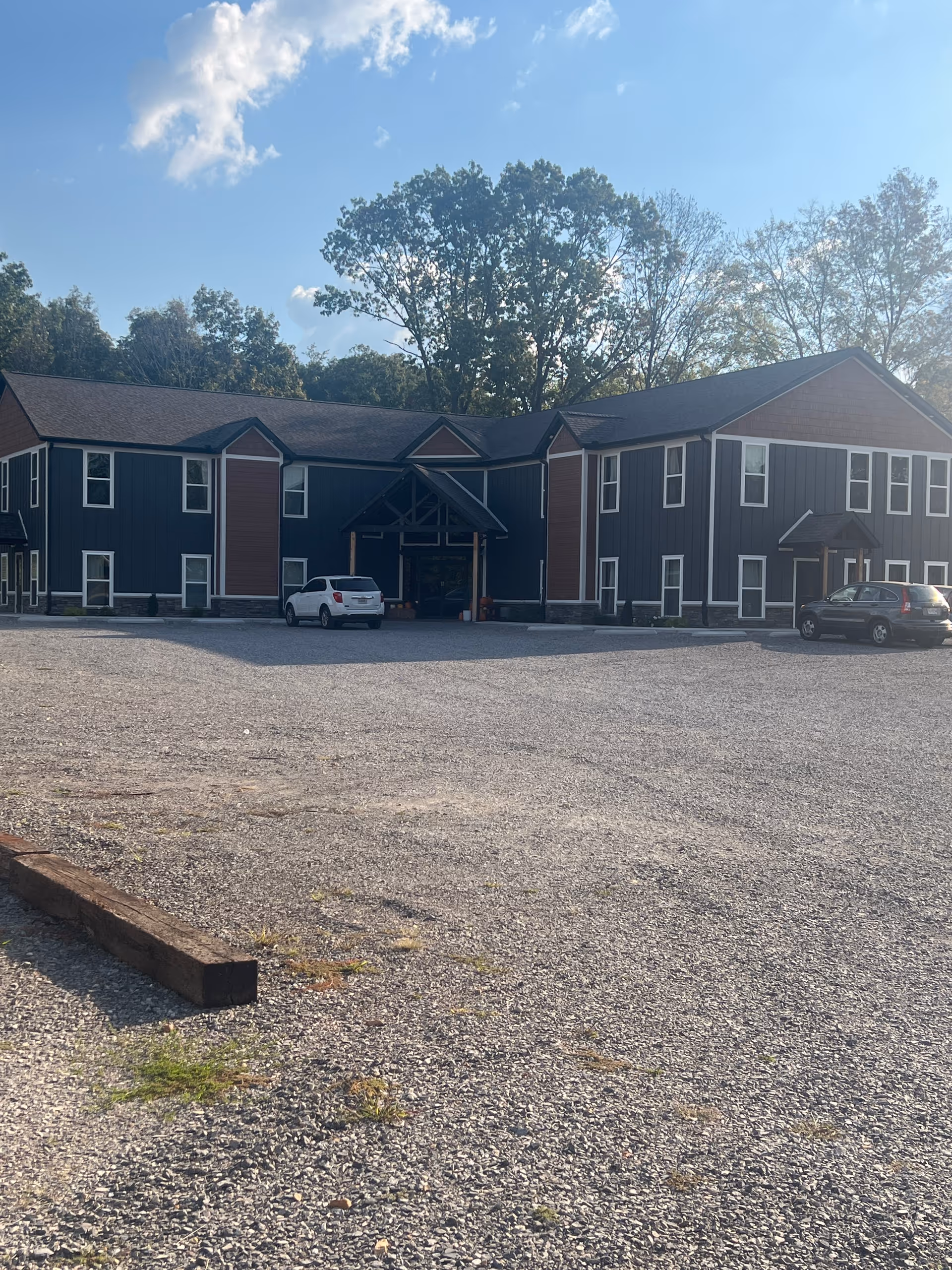 Two-story blue building with peaked entrances, gravel parking lot, and trees behind.
