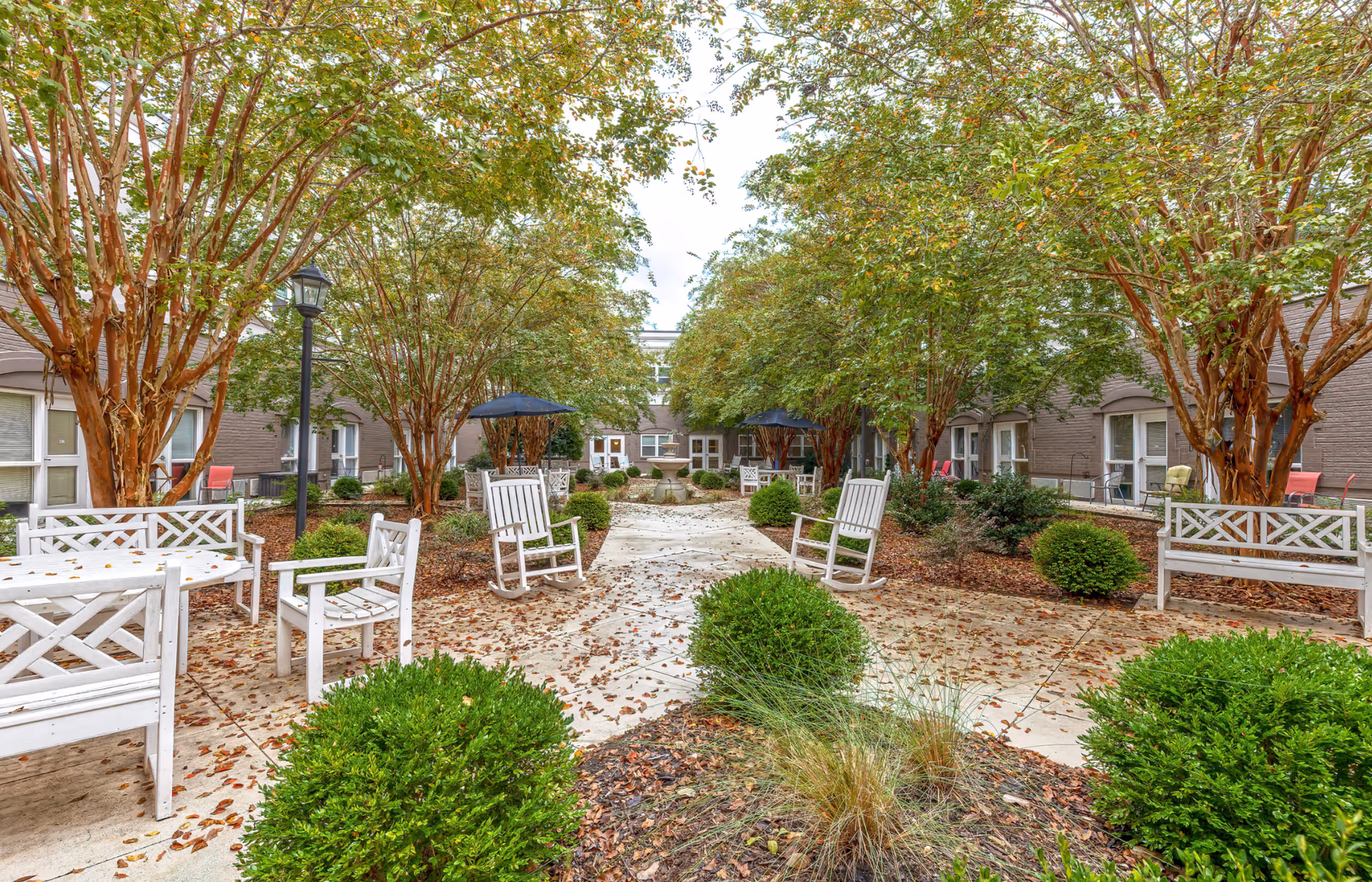 Outdoor courtyard area at The Inverness at Spartanburg featuring a paved walkway lined with green bushes and trees. White wooden benches, chairs, and rocking chairs are arranged along the path, with black umbrellas providing shade over some seating areas. The courtyard is surrounded by the building with windows and doors visible.