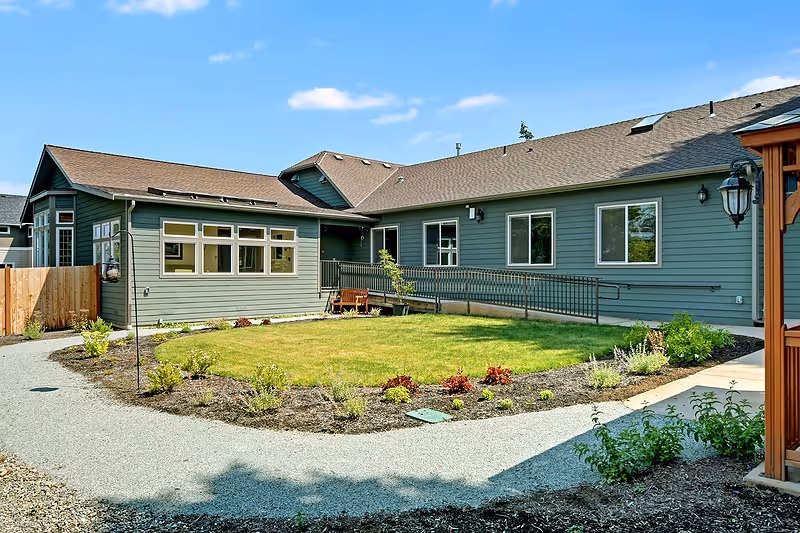 Exterior view of a single-story building with green siding and a brown roof surrounding a small courtyard with a grassy area and landscaped plants. There is a paved pathway around the courtyard and a wooden fence on the left side. The sky is clear and blue.