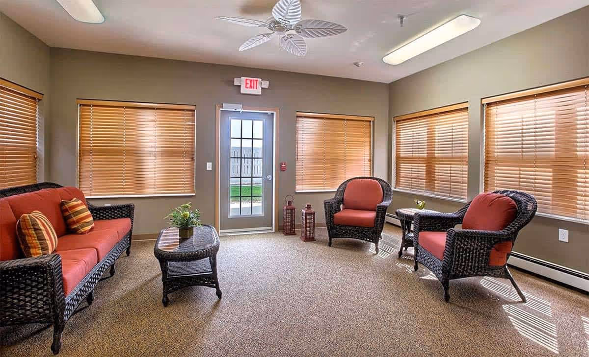 A cozy indoor lounge with wicker seating, red cushions, and blinds in a senior living facility.