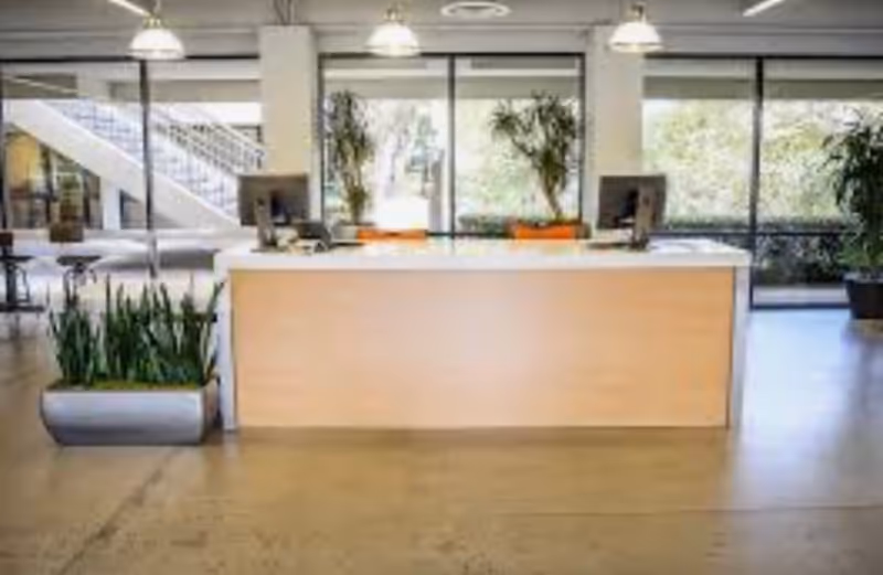 Reception desk in a bright lobby with plants and large windows inside a modern facility.