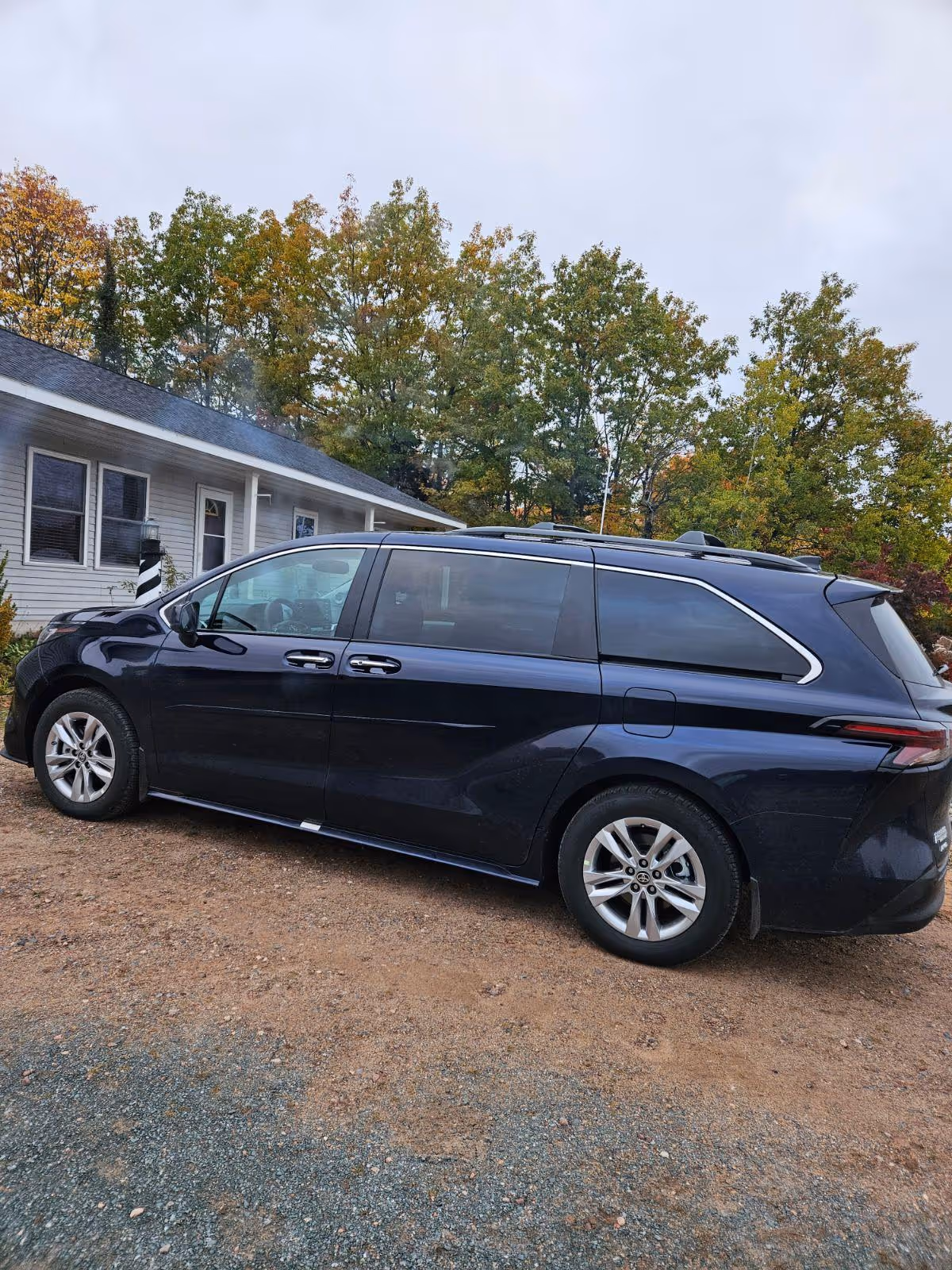 A dark minivan parked on a dirt lot in front of a light building with trees.