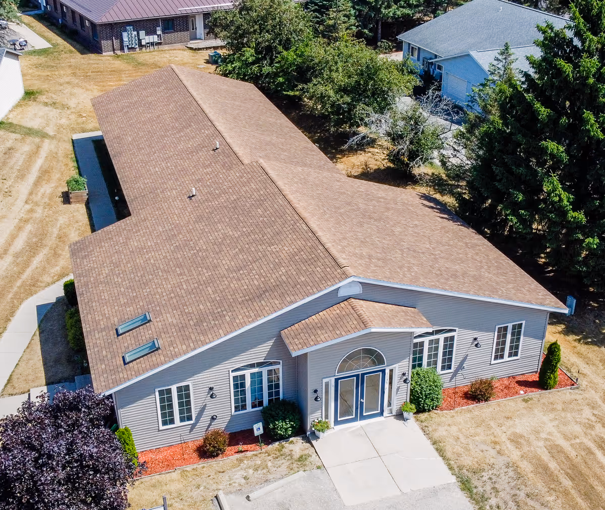 Aerial view of a single-story building with a brown shingled roof and light gray siding. The building has a main entrance with double doors and an arched window above. There are several windows on the visible sides, surrounded by small bushes and landscaping with red mulch. The surrounding area includes a driveway, a sidewalk, and some trees and grass.