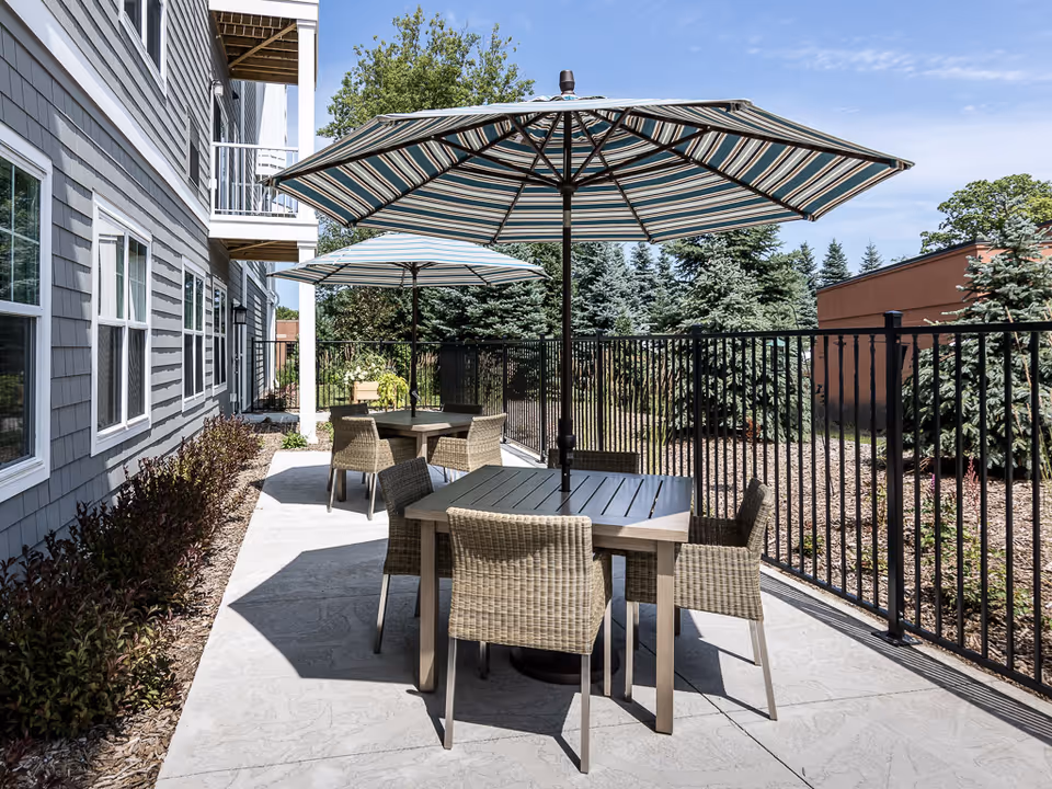 Outdoor patio area with two tables, each shaded by a large striped umbrella. Each table is surrounded by four wicker chairs. The patio is adjacent to a building with gray siding and white-framed windows, and is bordered by a black metal fence with trees and landscaping beyond.