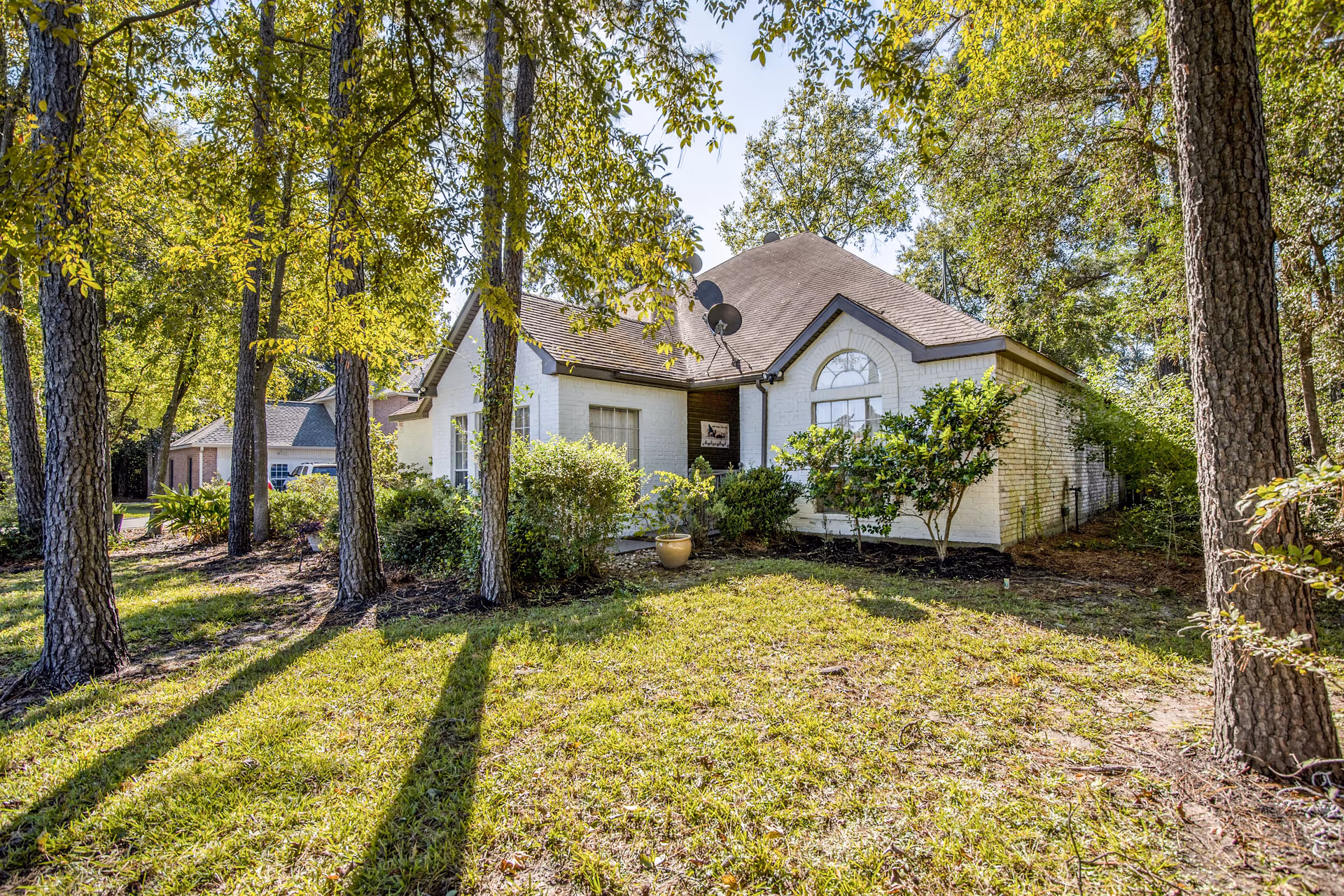A single-story white brick house with a brown shingled roof surrounded by trees and greenery on a sunny day. The house has a large arched window and a satellite dish on the roof. The lawn is grassy with some bushes and plants near the house.