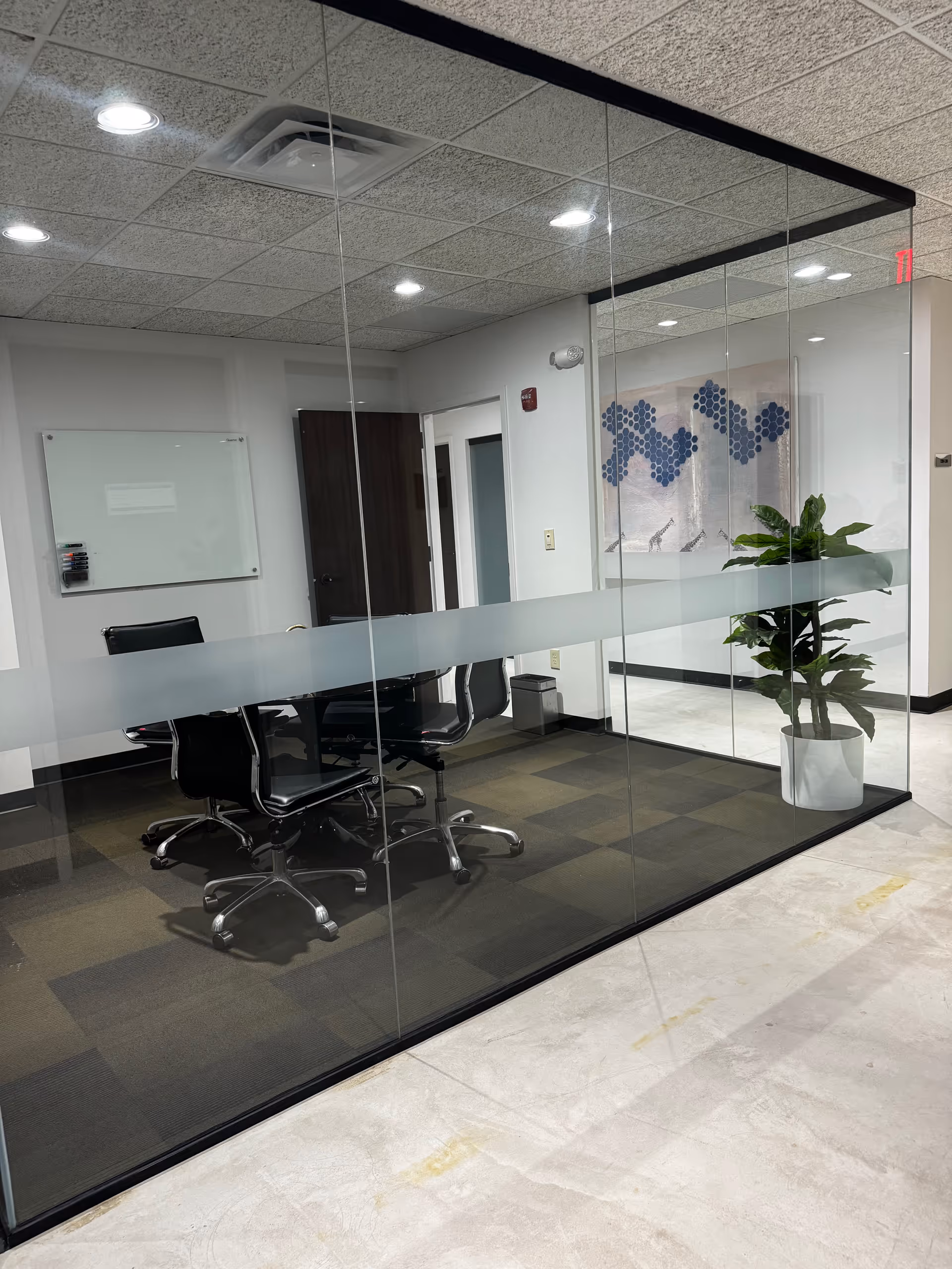 Glass-walled conference room with a table, chairs, whiteboard, and a potted plant.