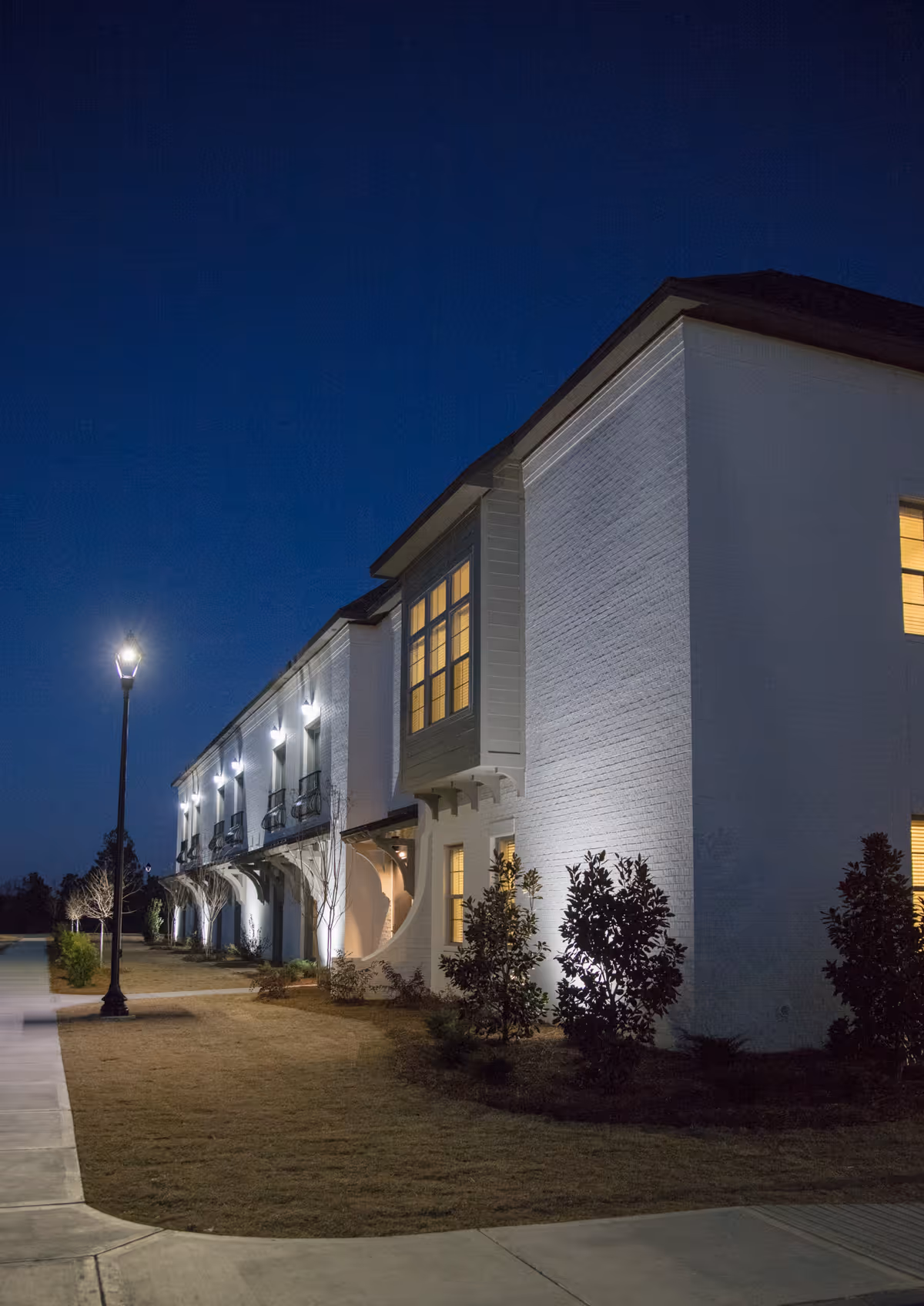 Exterior view of a two-story white brick building at night with illuminated windows and outdoor lighting along a sidewalk and landscaped area with small trees and bushes.