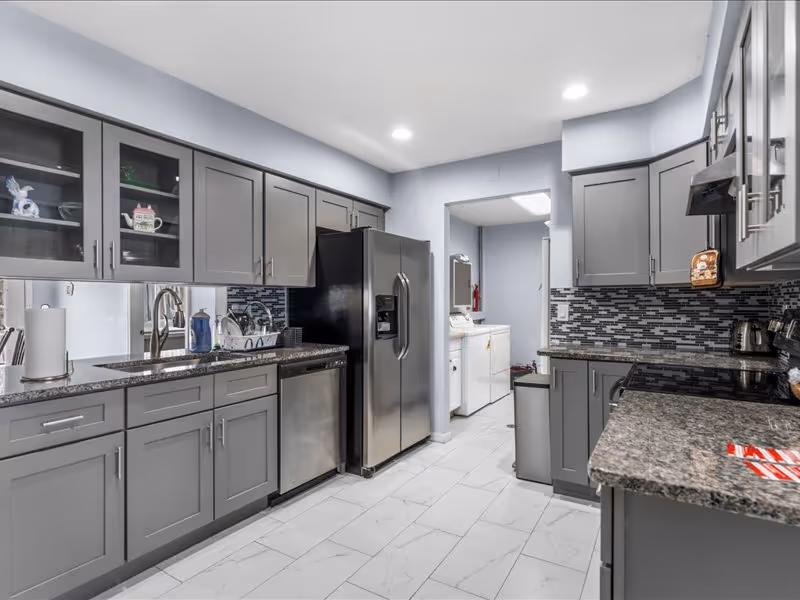 Modern kitchen with gray cabinets, granite countertops, stainless steel refrigerator, dishwasher, and stove. The kitchen has a tiled floor and a mosaic tile backsplash. There is a doorway leading to a laundry area with a washer and dryer visible.