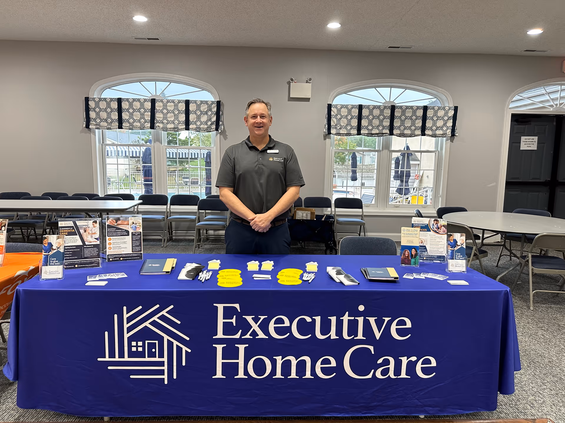 A staff member stands behind a blue Executive Home Care table at an indoor event.
