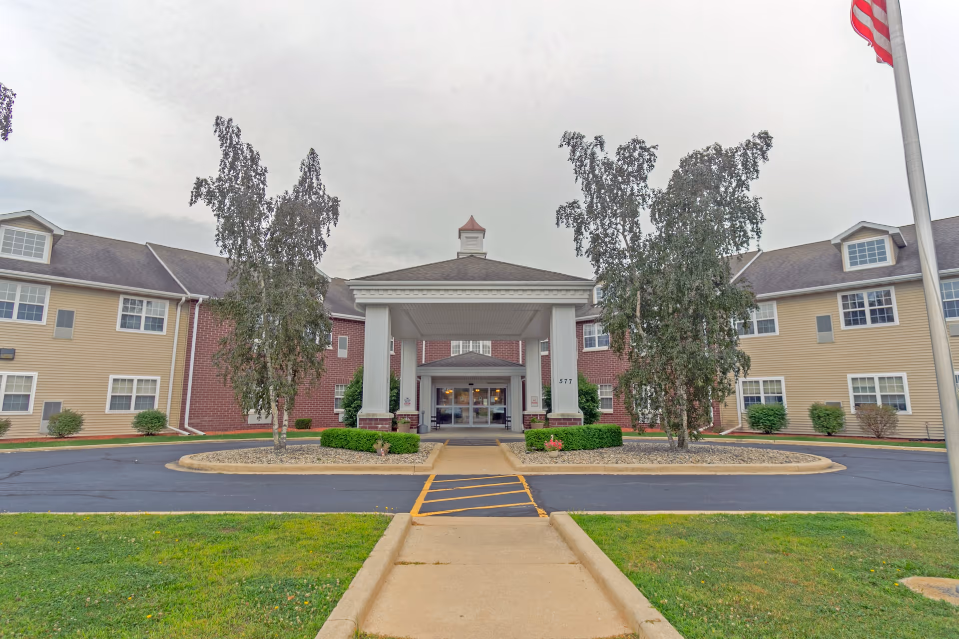Front exterior view of Heritage Woods of Watseka, showing a large building with a covered entrance supported by white columns, two trees in front, a paved driveway, and an American flag on a pole to the right.