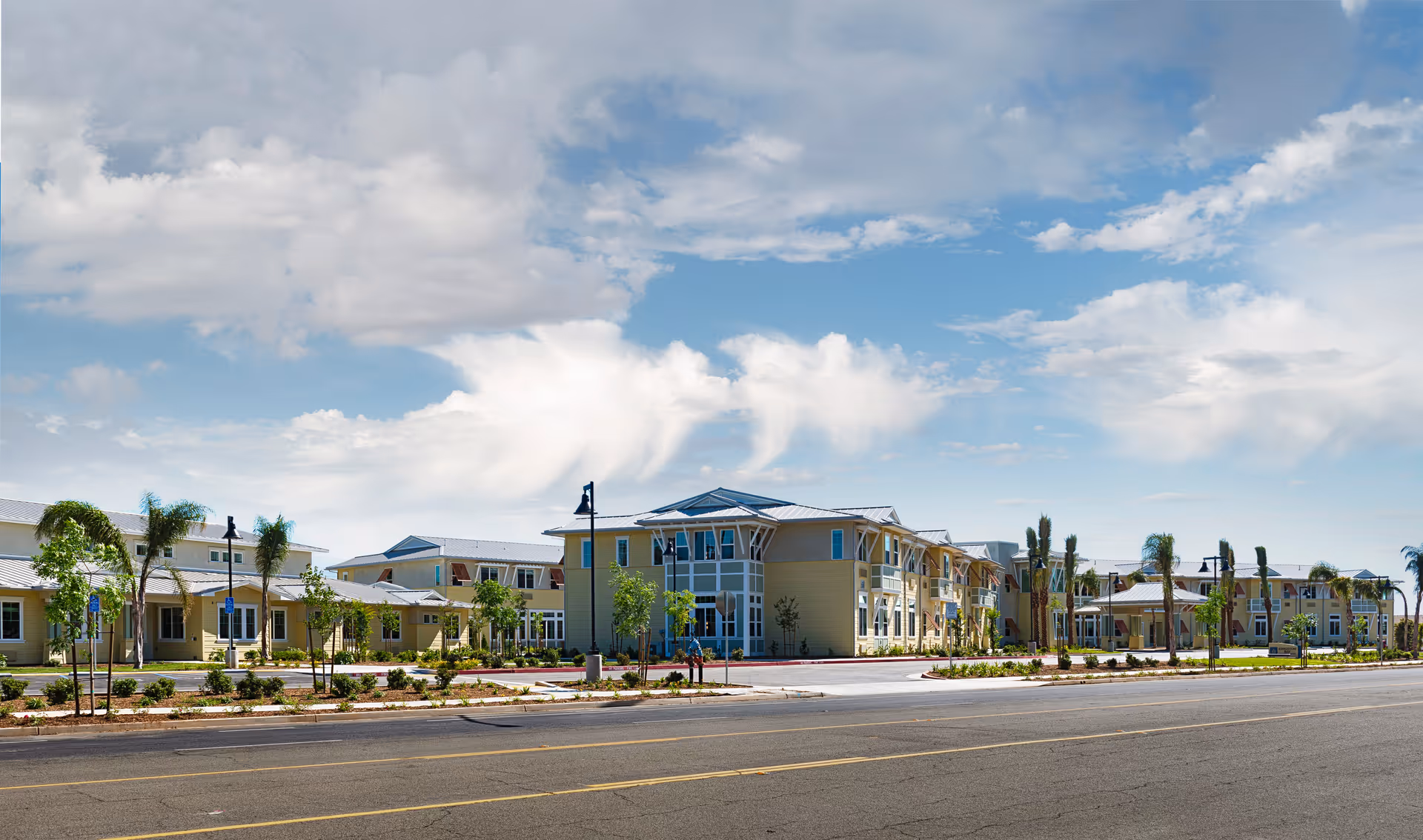 Front view of a low-rise senior living complex with yellow buildings, palm trees, landscaped grounds and a road in the foreground under a blue sky.