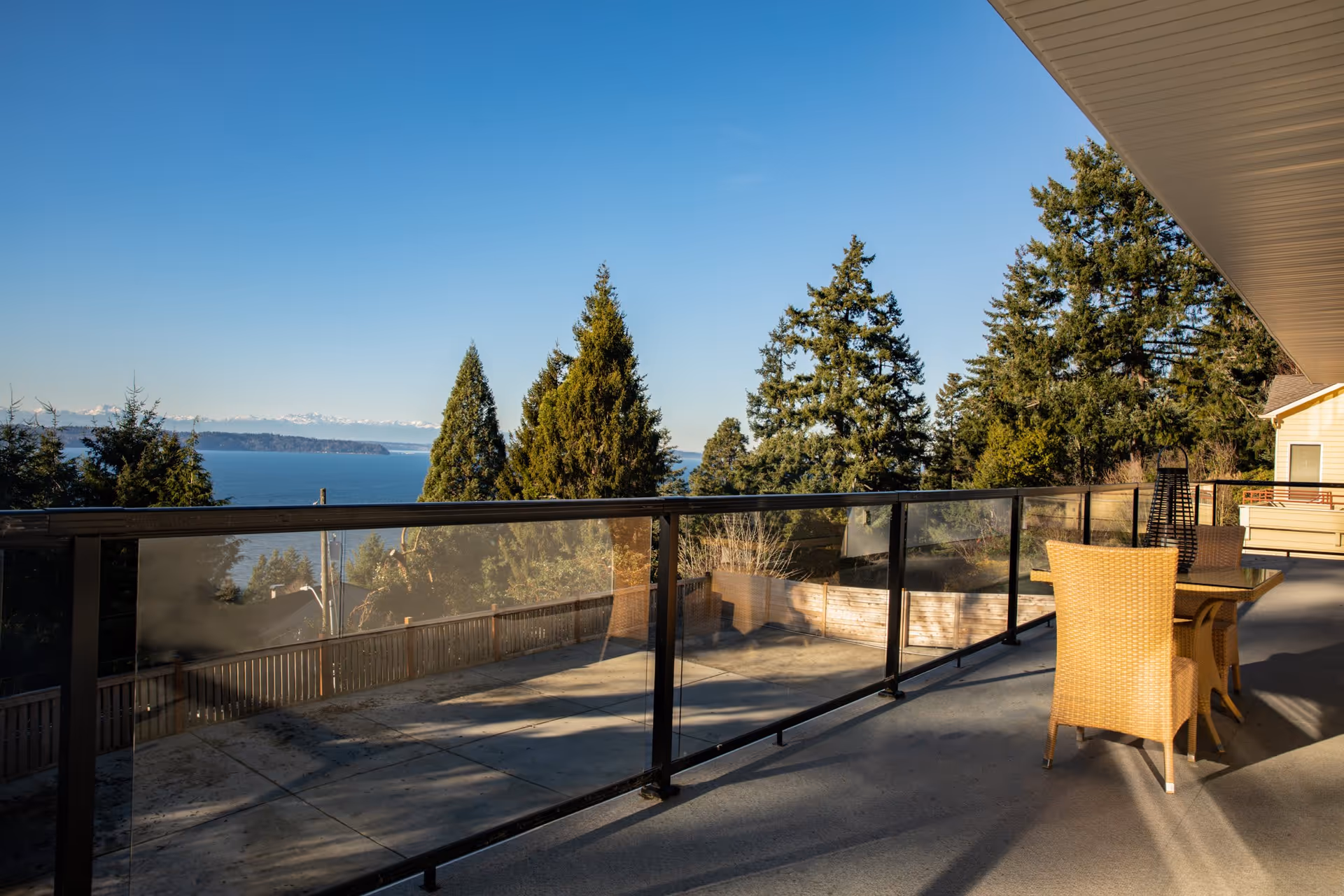 Outdoor patio area with a glass railing overlooking a scenic view of trees, a body of water, and distant mountains under a clear blue sky. The patio has a table with two wicker chairs and a tall outdoor heater.