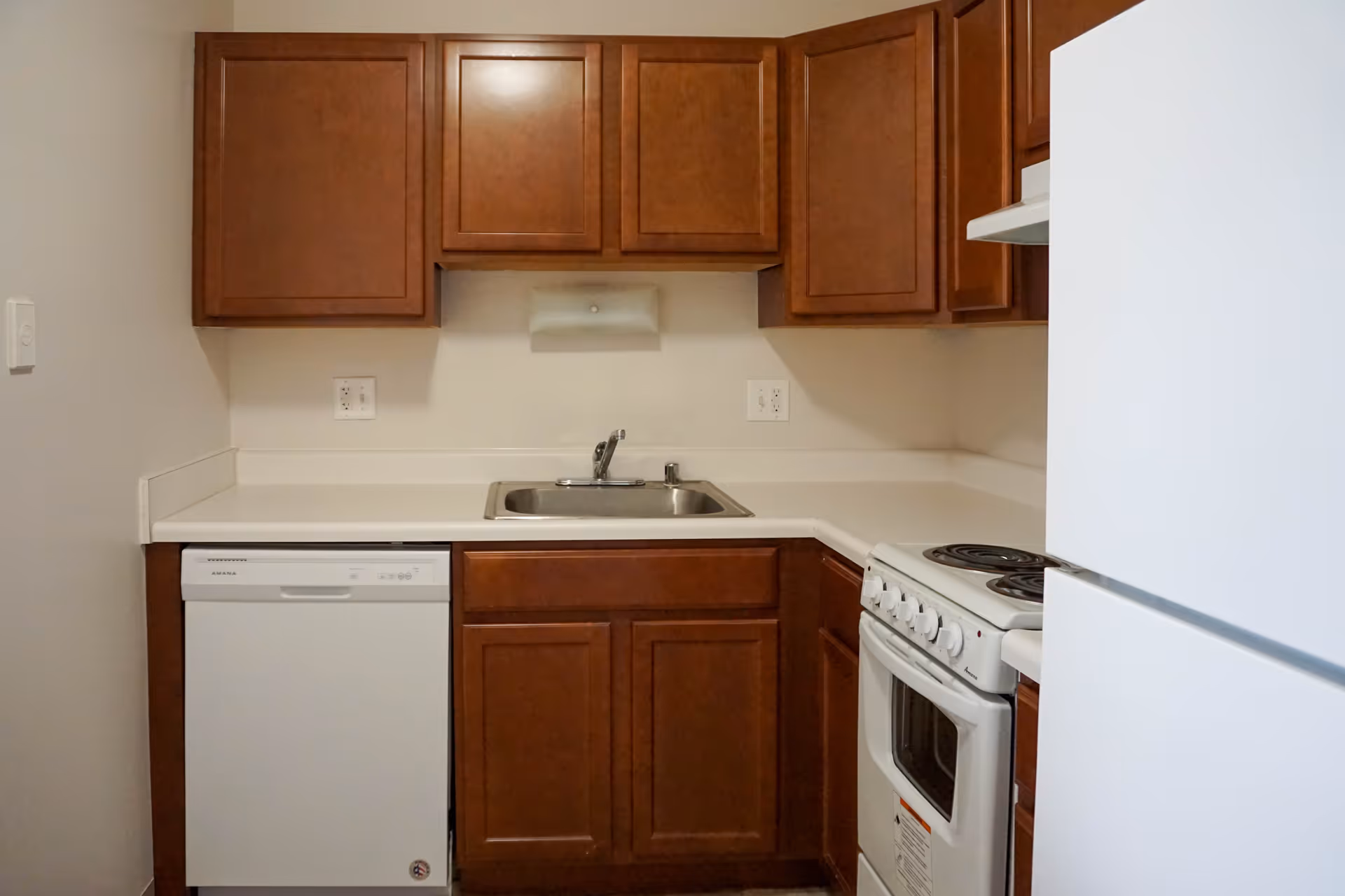Compact kitchen with wooden cabinets, sink, dishwasher, and a white stove.
