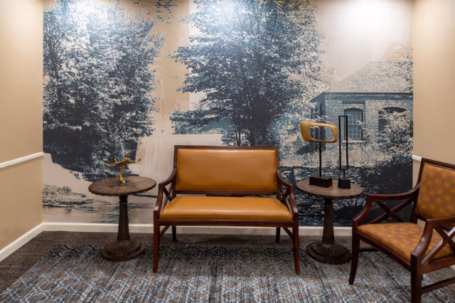 A seating area with a brown leather loveseat and a matching armchair with patterned upholstery. Two round wooden side tables flank the loveseat, one with a small decorative airplane and the other with two modern sculptures. The background features a large mural depicting a scenic outdoor scene with trees, a waterfall, and a building.