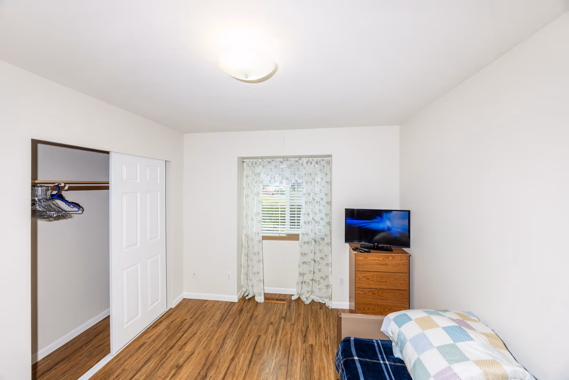 A small bedroom with a window, closet, dresser, and bed in a senior living facility.