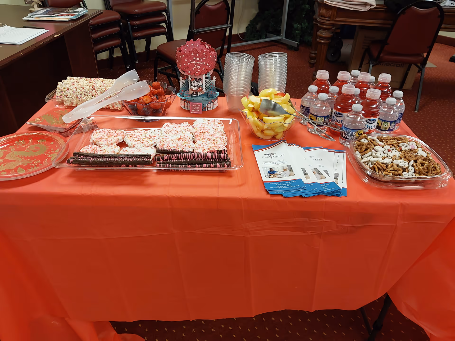 Table with red tablecloth displaying cupcakes, fruit, pretzels, water bottles, and brochures.