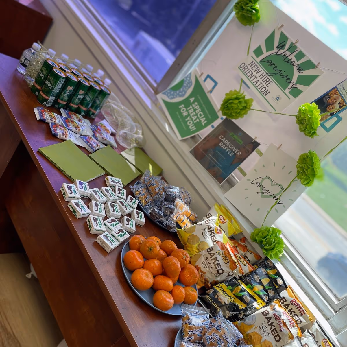 Snack table with oranges, bags of snacks, juice boxes, and green decorations in an indoor facility.