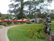 Curved brick walkway through a grassy garden with statues and a row of vintage red cars parked along the driveway under trees.
