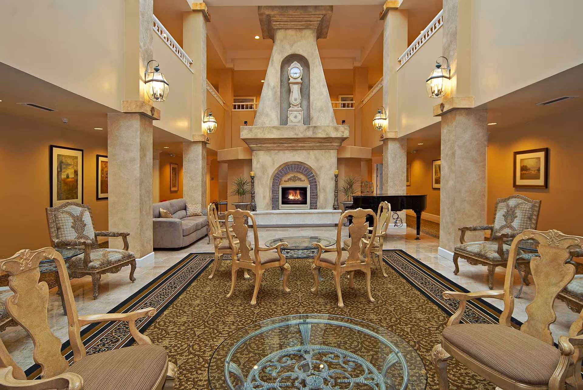 A spacious and elegant living room area with a large stone fireplace as the focal point. The room features multiple ornate wooden chairs with cushions arranged around glass-top tables on a patterned carpet. There are two gray sofas along the walls, framed artwork, wall-mounted lantern-style lights, and a black grand piano in the background. The room has high ceilings with a balcony railing visible on the upper level.