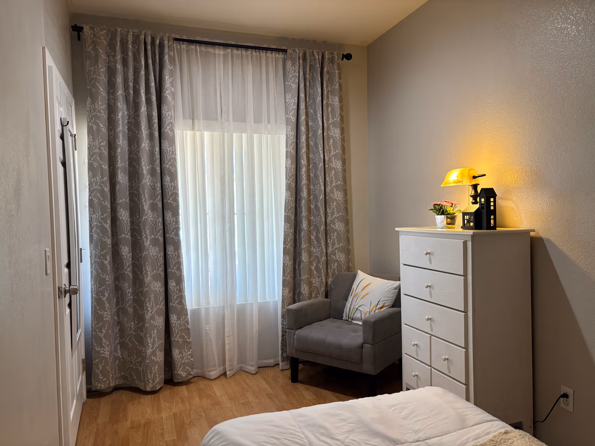 A small cozy bedroom with a dresser, chair, bed, and large window with patterned curtains.