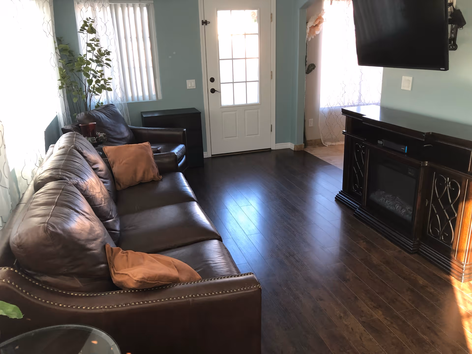 Sunlit living room with a brown leather sofa and pillows, a wall-mounted TV over a wooden media console, and a white door with windows at the far end.