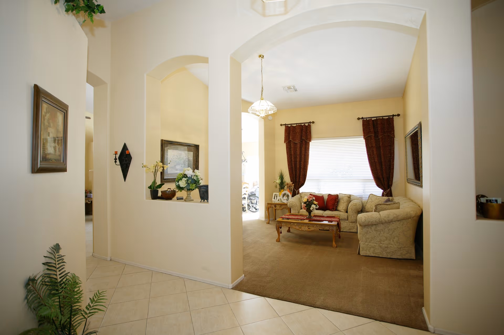 Interior view of a living room area with beige walls and carpet. The room features a beige patterned sofa and armchair with red cushions, a wooden coffee table with a floral centerpiece, and a side table with framed photos and plants. Large windows with dark red curtains allow natural light to fill the space. The foreground shows a tiled floor hallway with decorative plants and wall art.