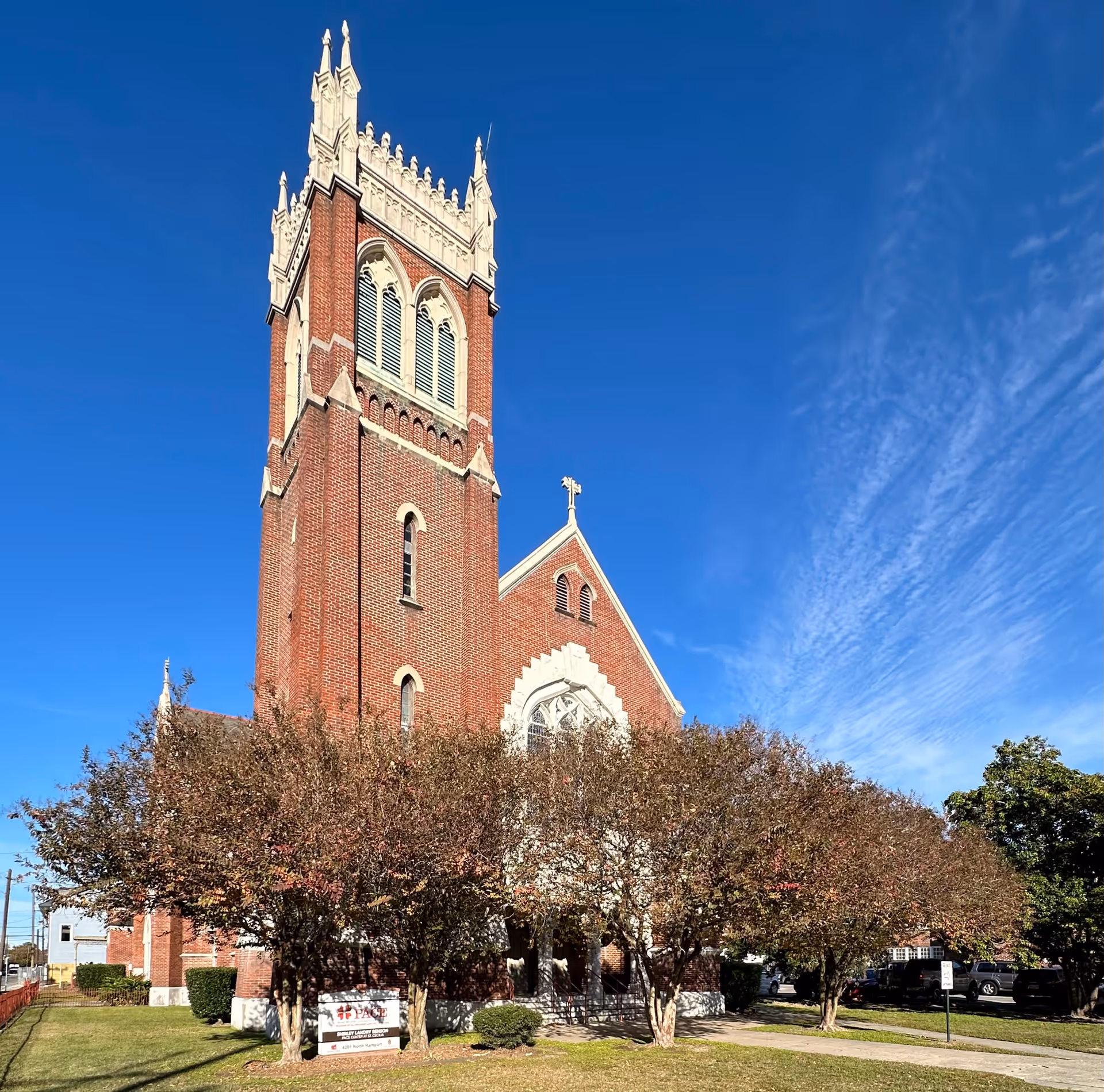 Red brick church with tall tower, arched windows, and surrounding trees under a blue sky.