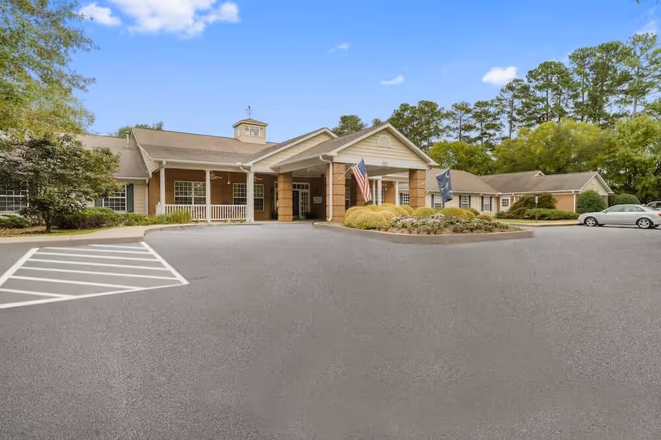Exterior front view of a single-story assisted living facility building with a covered entrance supported by brick columns, an American flag, and a landscaped area with bushes and flowers. The building is surrounded by trees and a clear blue sky.