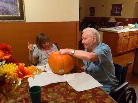 An elderly man and woman sitting at a table indoors, carving a large pumpkin together. The man is in a wheelchair and is actively working on the pumpkin, while the woman watches and smiles. The table is covered with a fall-themed tablecloth and some flowers are visible in the foreground. The background shows a kitchen area with cabinets and a counter.