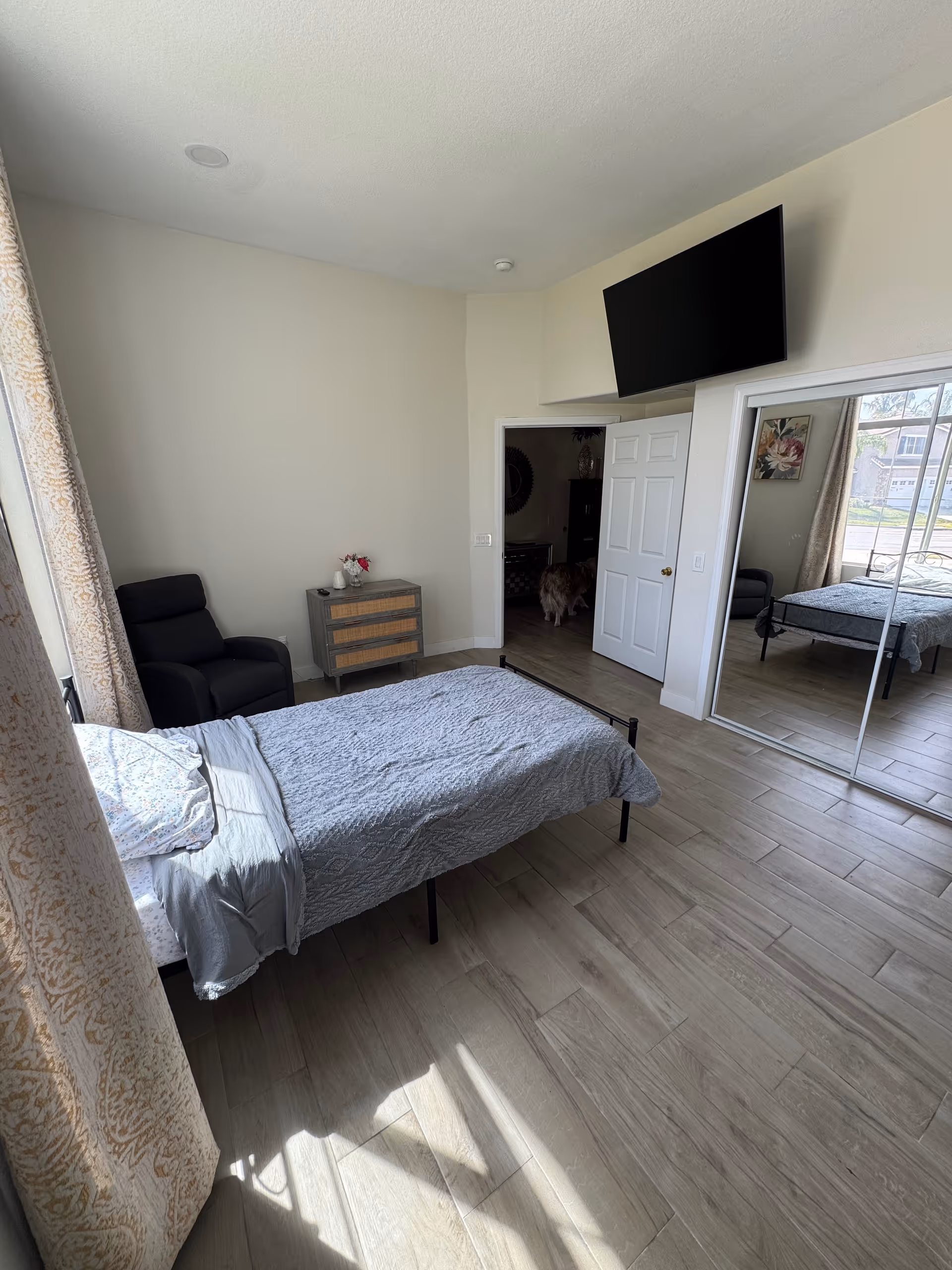 Bright bedroom with a bed, dresser, chair, mounted TV, and mirrored closet doors.