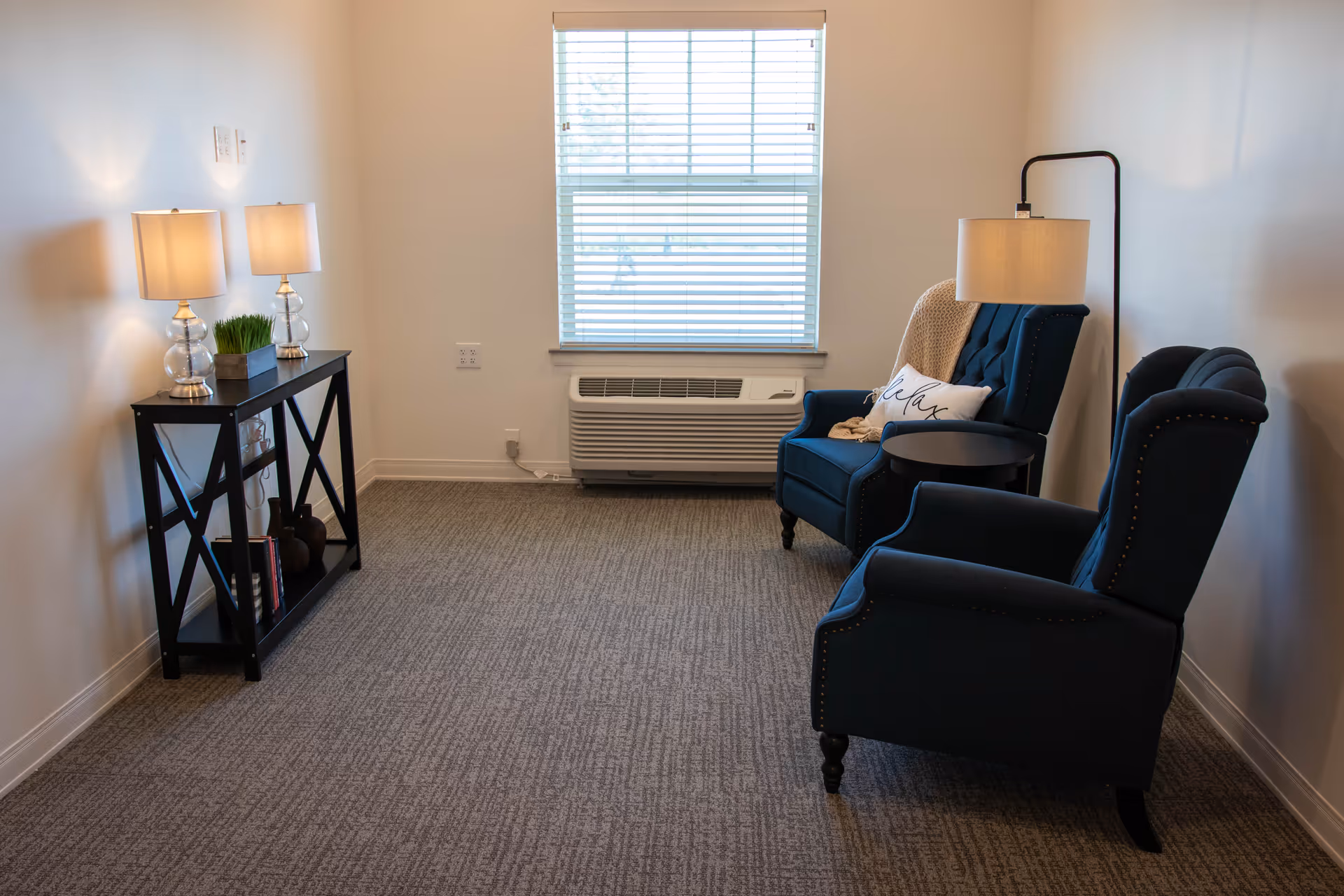 Cozy lounge area with blue armchairs, a side table, lamps, and a window air conditioner.
