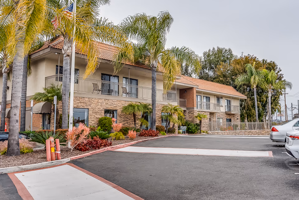 Exterior view of a two-story senior living facility with a tiled roof, balconies, palm trees, and landscaped bushes. There is a parking lot in the foreground with several parked cars and a flagpole with an American flag.