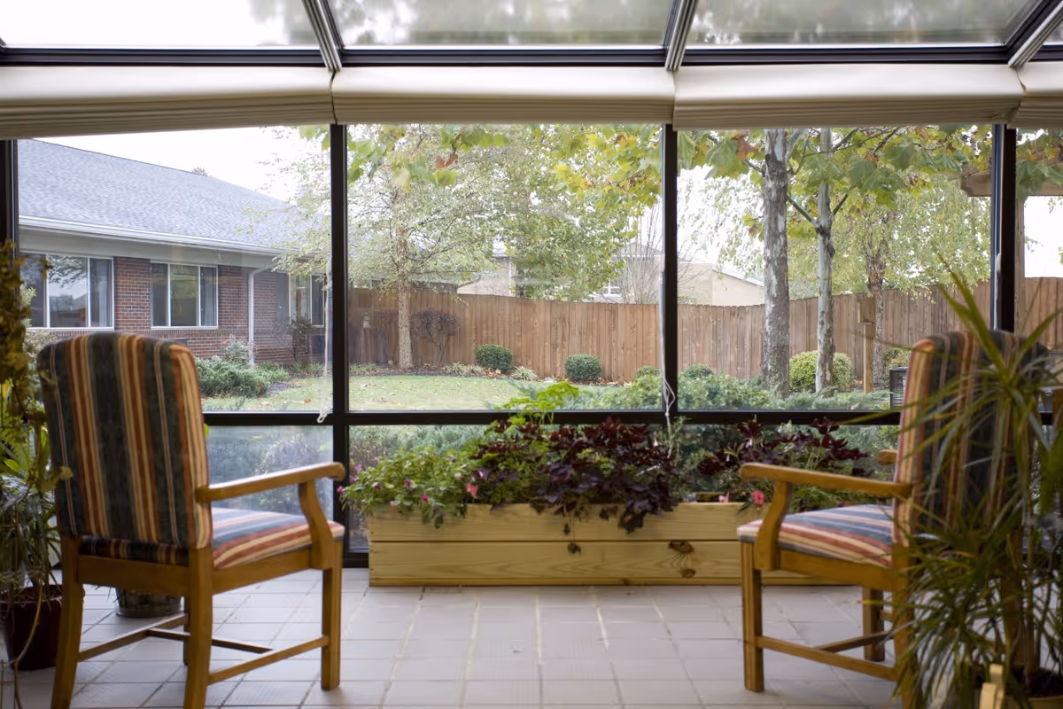 Two wooden chairs with striped cushions facing each other in a sunroom with large glass windows overlooking a fenced garden with trees and shrubs.