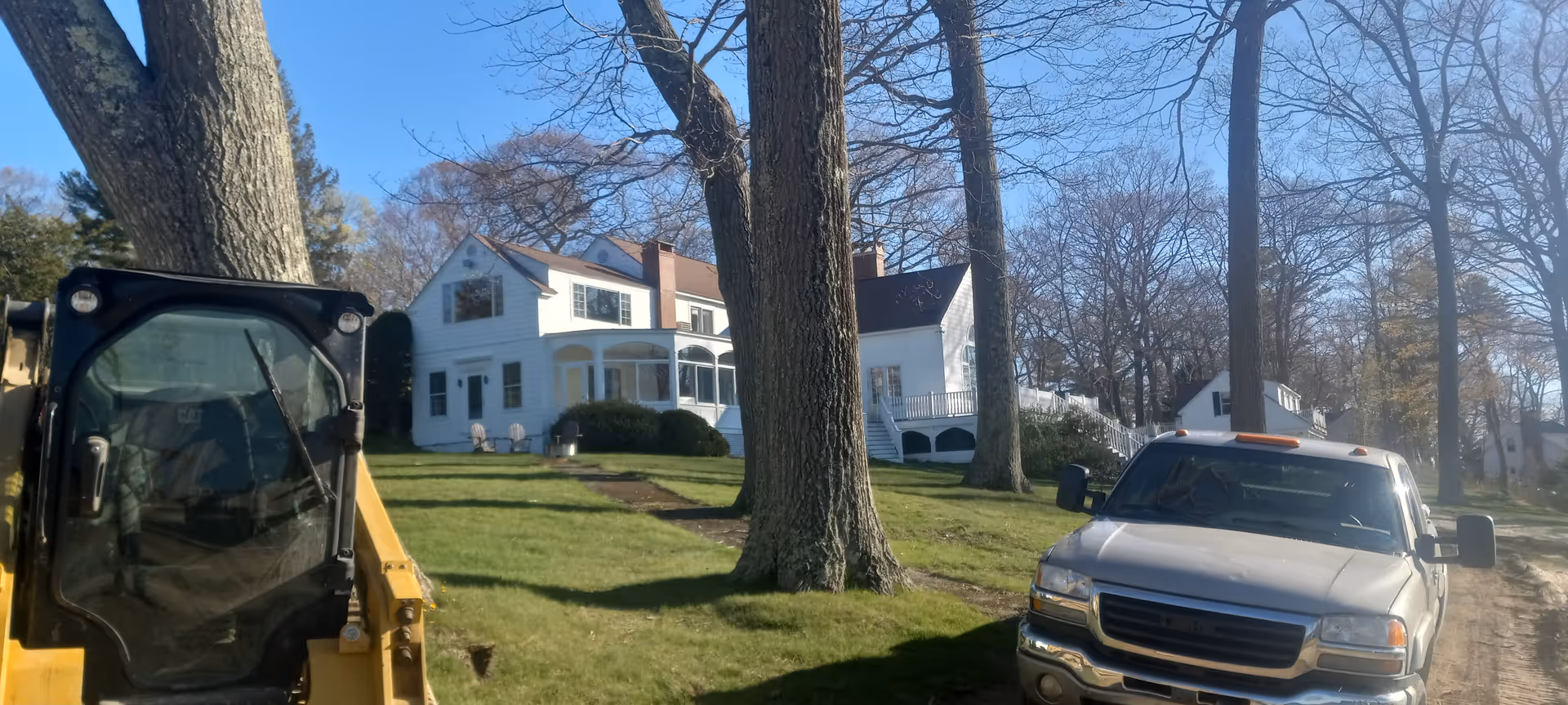 Sunlit exterior of a white two-story house with trees, a yellow excavator, and a white truck.